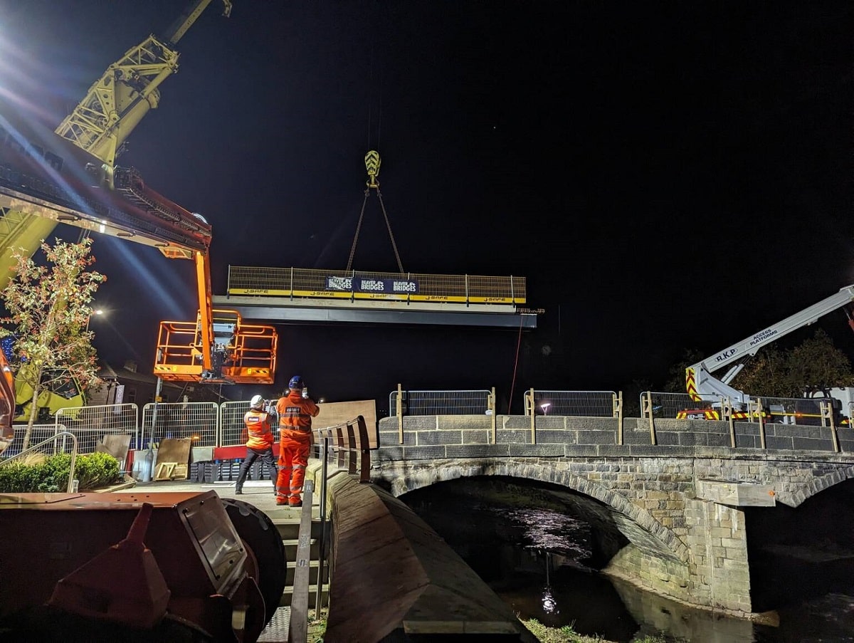 COnstruction workers guide a crane lifting bridge beams into place in Mytholmroyd at night.