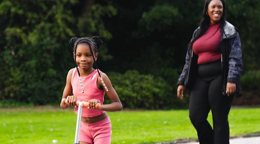 Woman walking and child scooting through a park.